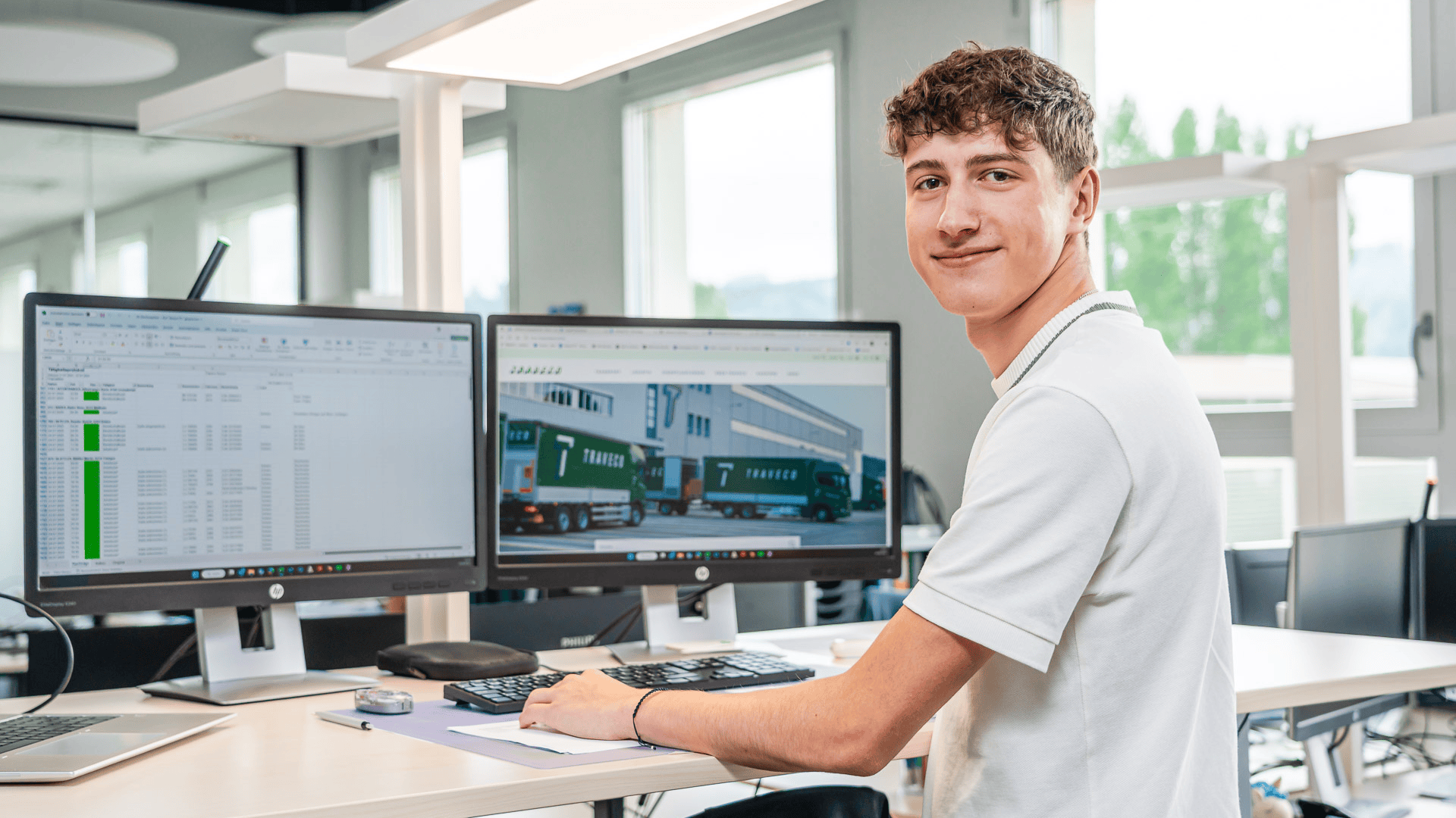 Ein junger Mann in einem weißen Hemd sitzt an einem Schreibtisch, lächelt in die Kamera und arbeitet an einem Computer mit zwei Monitoren, auf denen Tabellen und Bilder von Lastwagen zu sehen sind – in einem hellen Büro.