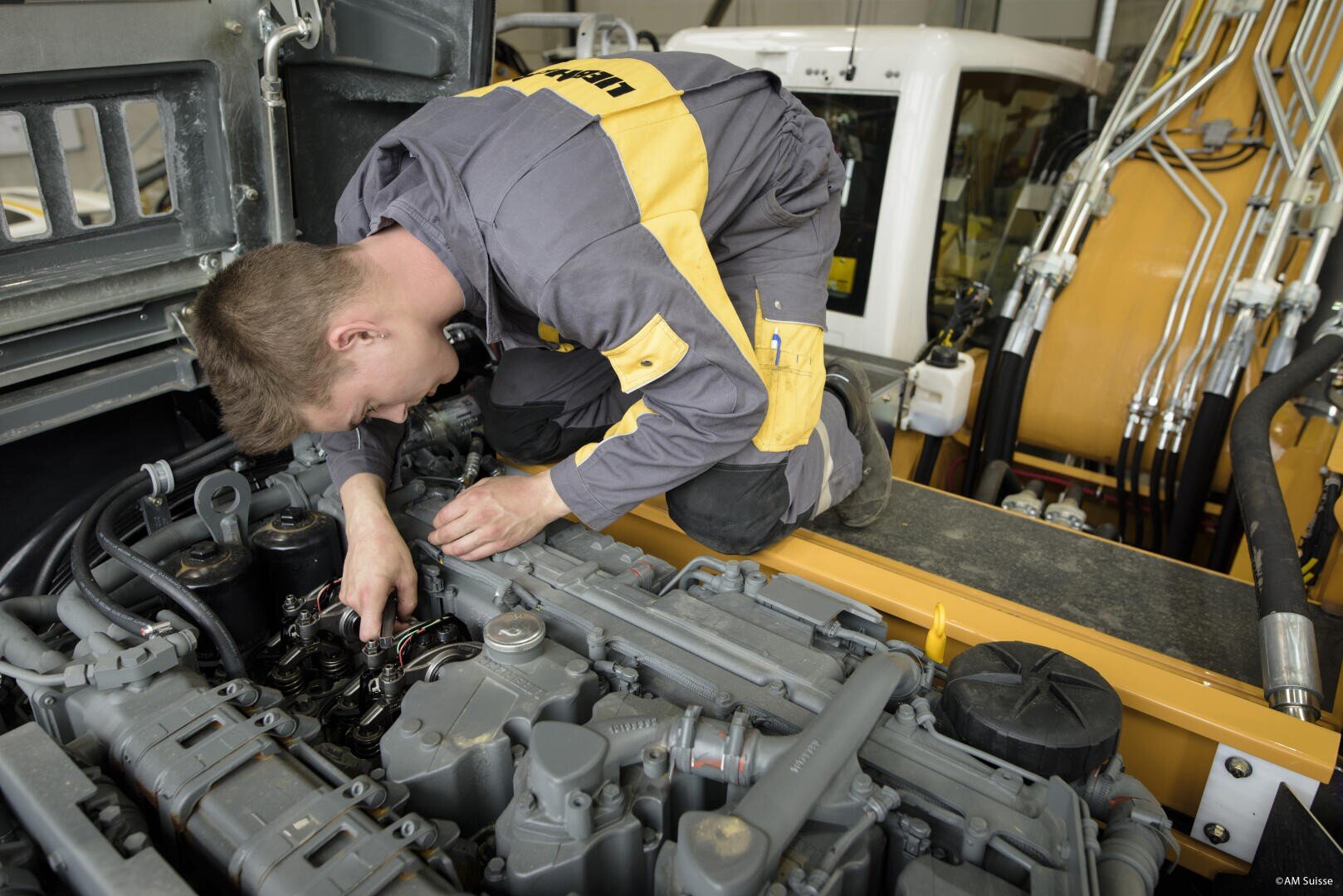 Ein Mechaniker in gelber und grauer Uniform kniet an einer großen Maschine, die er in einer Werkstatt oder Garage inspiziert und an deren Motor er mit großer Aufmerksamkeit arbeitet.