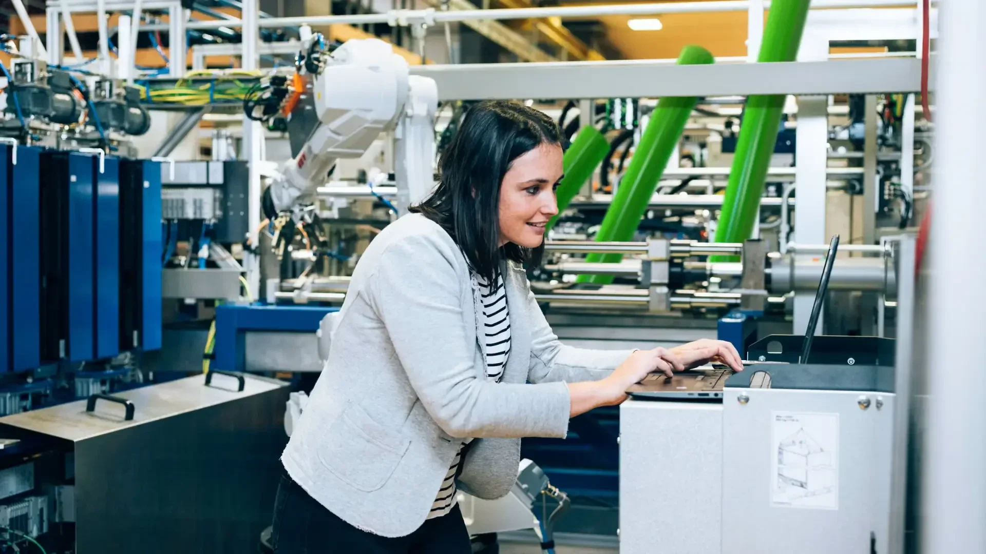 Eine Frau in einem hellen Blazer arbeitet mit Maschinen in einer modernen Industrieumgebung und tippt auf einem Bedienfeld, umgeben von Geräten und Roboterarmen.