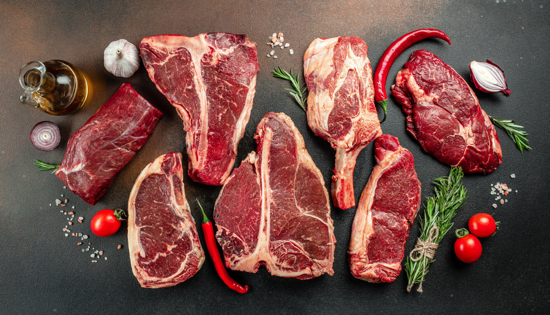 Verschiedene rohe Rindersteaks auf einem dunklen Untergrund, umgeben von Kirschtomaten, roten Zwiebeln, Knoblauch, Chilischoten, Rosmarin und einer Flasche Öl.