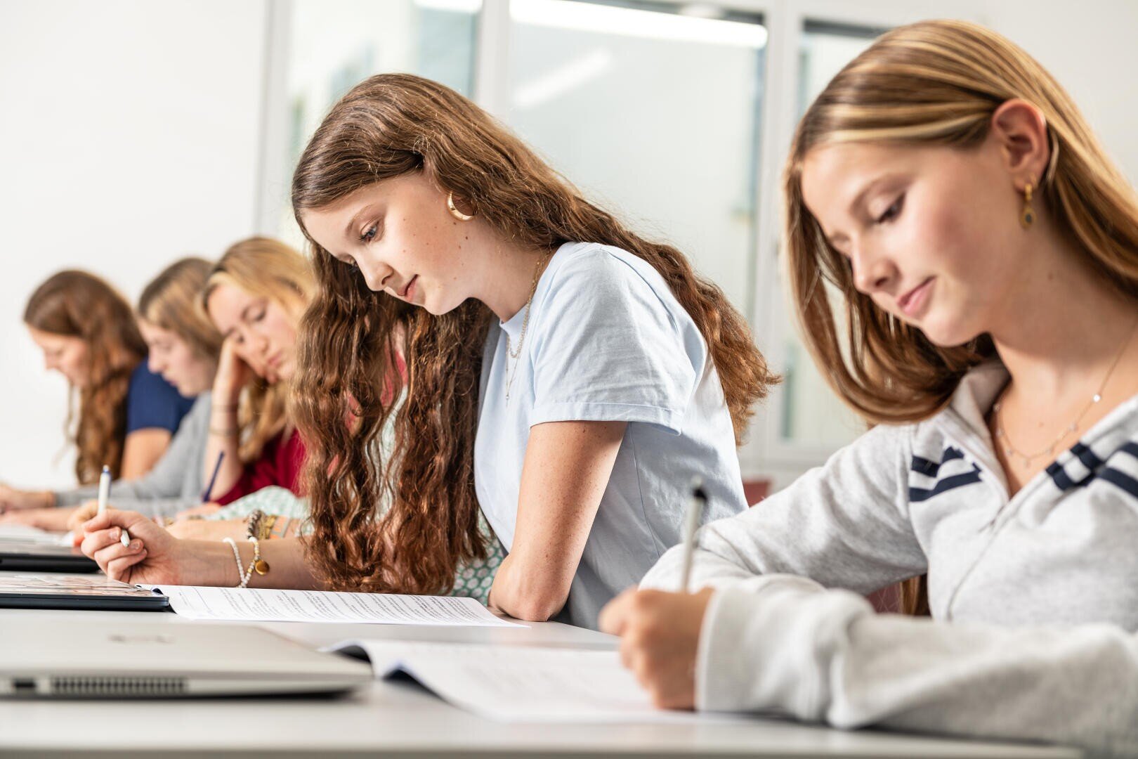Eine Gruppe Jugendlicher sitzt an den Tischen in einem Klassenzimmer und schreibt in Hefte. Sie scheinen konzentriert und mit ihrer Arbeit beschäftigt zu sein. Auf den Tischen sind auch ein Laptop und Papiere zu sehen.