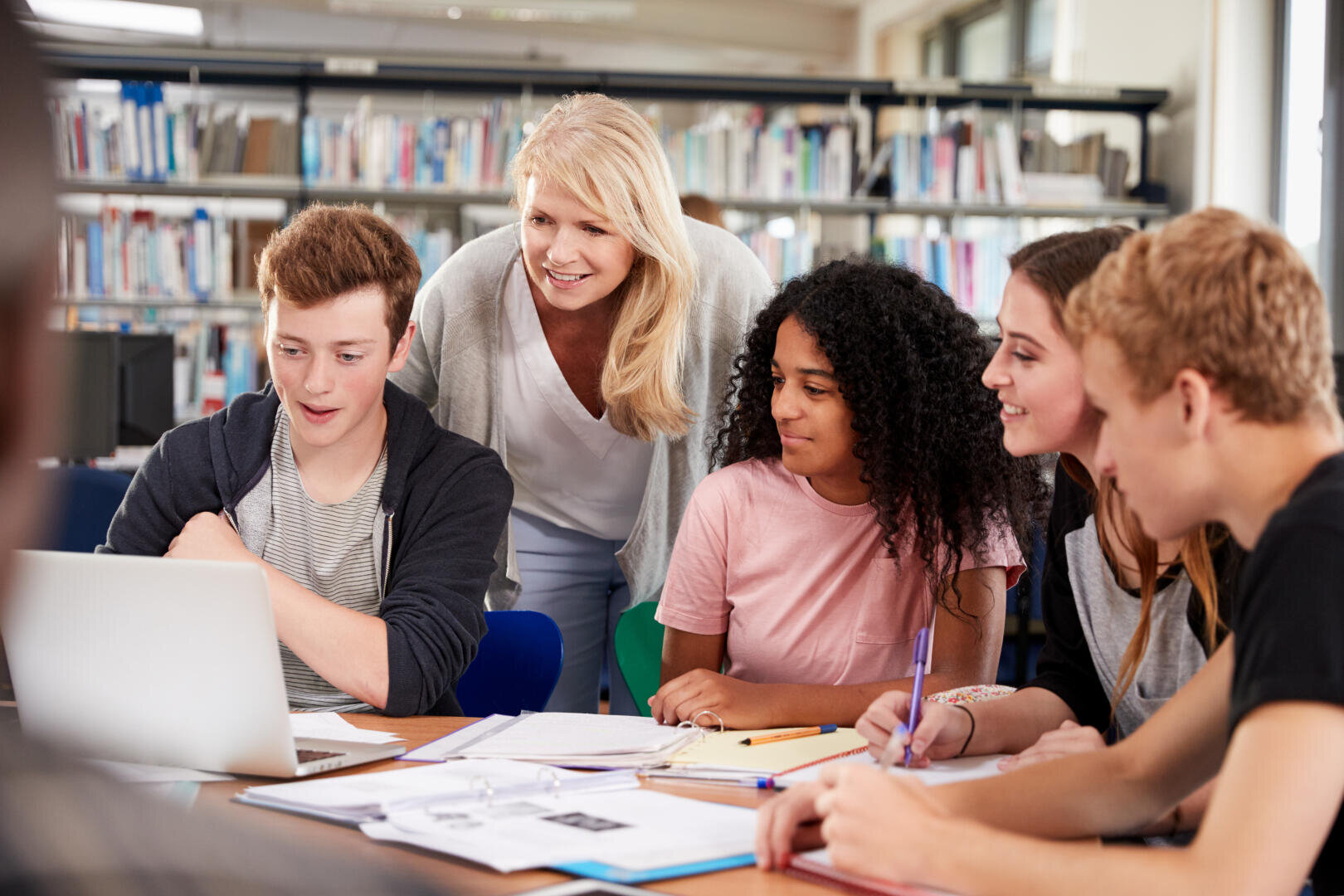 Eine Lehrerin steht lächelnd hinter einer Gruppe von vier Schülern, die an einem Tisch in einer Bibliothek sitzen und gemeinsam an einem Laptop arbeiten, vor dem Bücher und Papiere ausgebreitet sind.