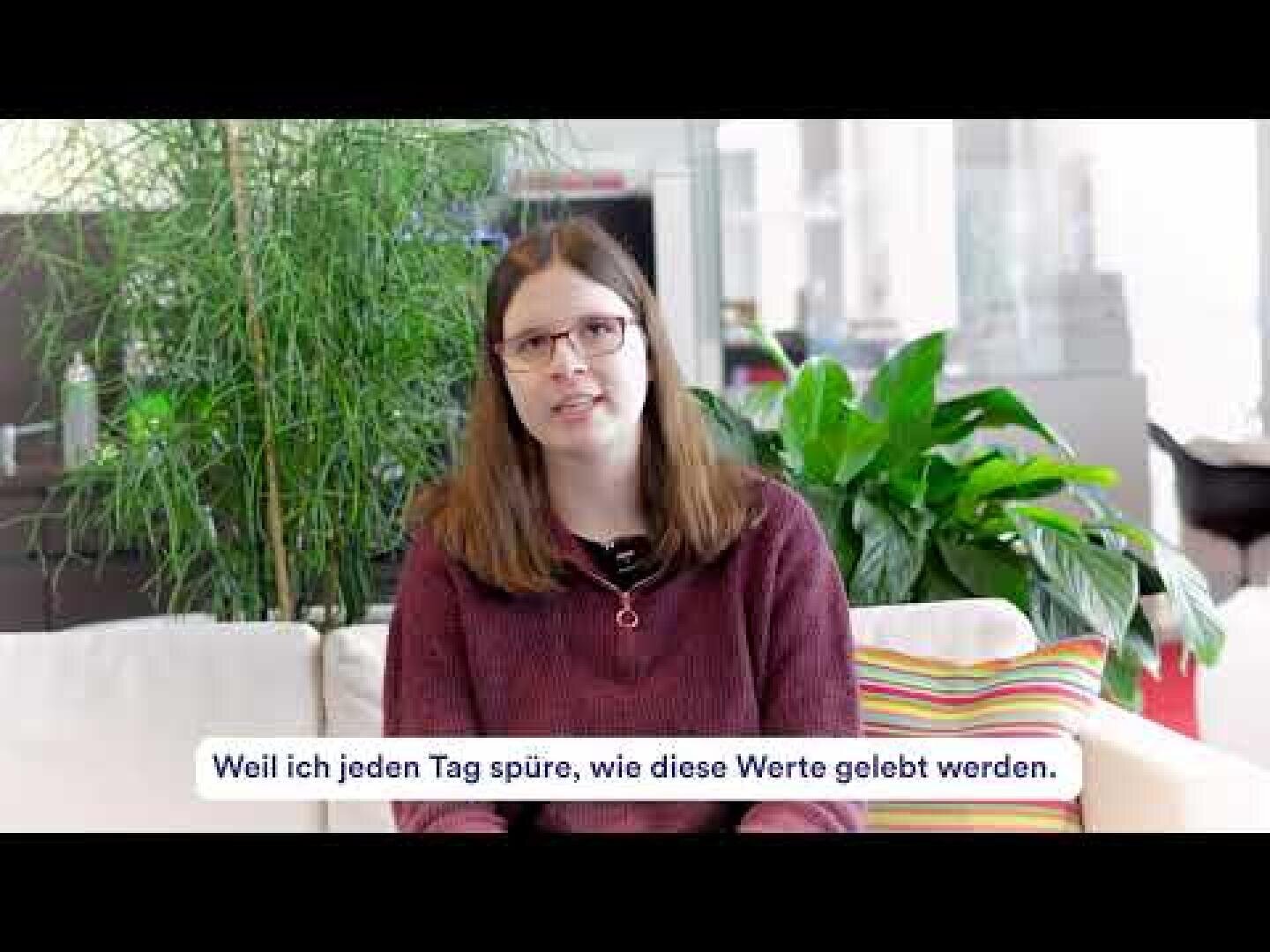 Eine Frau mit langen braunen Haaren und Brille sitzt auf einer weißen Couch mit bunten Kissen, im Hintergrund grüne Pflanzen. Der deutsche Text auf dem Bild lautet: Weil ich jeden Tag spüre, wie diese Werte gelebt werden.