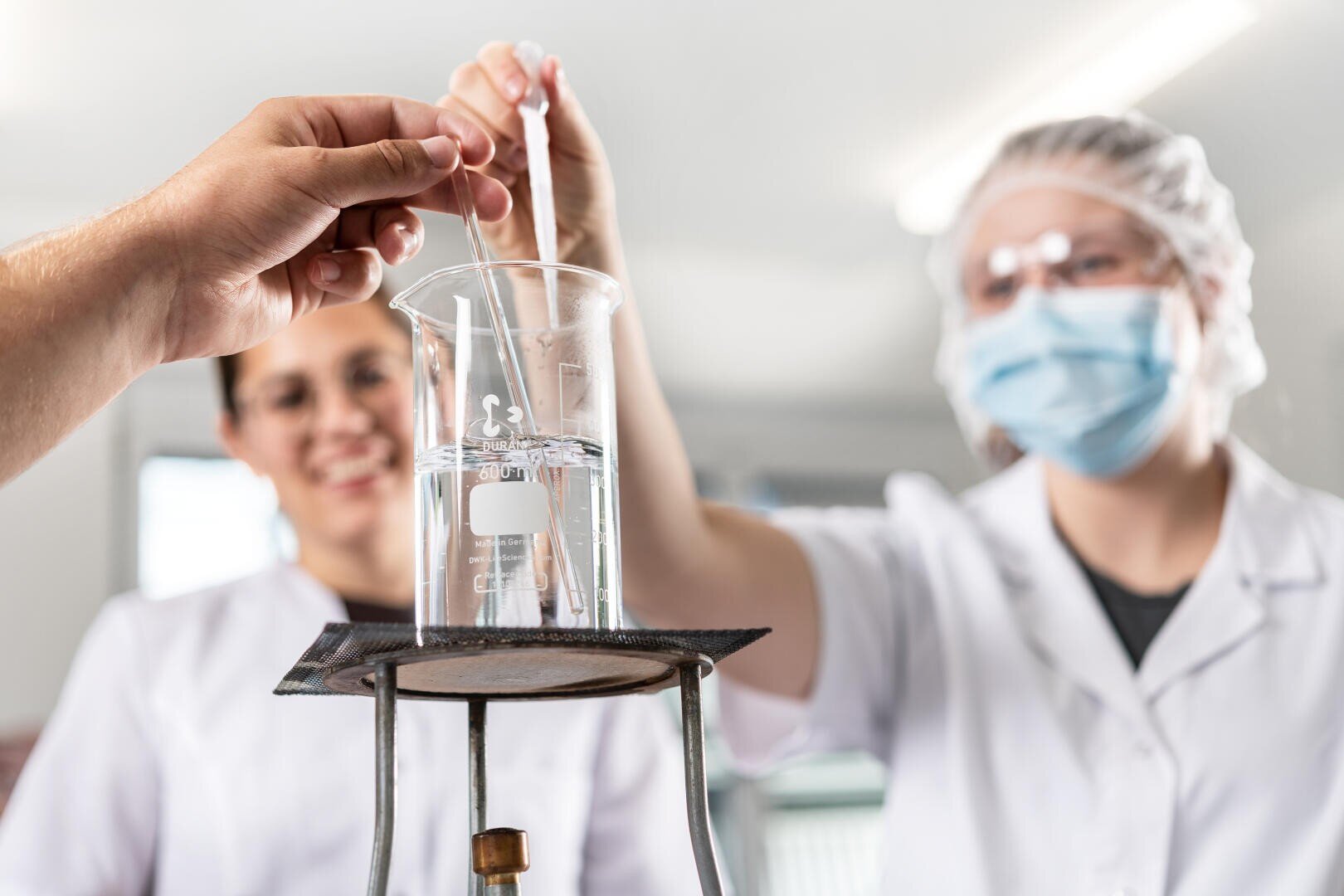 Zwei Personen in Laborkitteln und Sicherheitsausrüstung führen ein Experiment durch, bei dem sie mit einer Pipette Flüssigkeit in ein Becherglas mit klarer Lösung auf einem Ständer geben, wobei eine Person das Becherglas hält und beide konzentriert wirken.