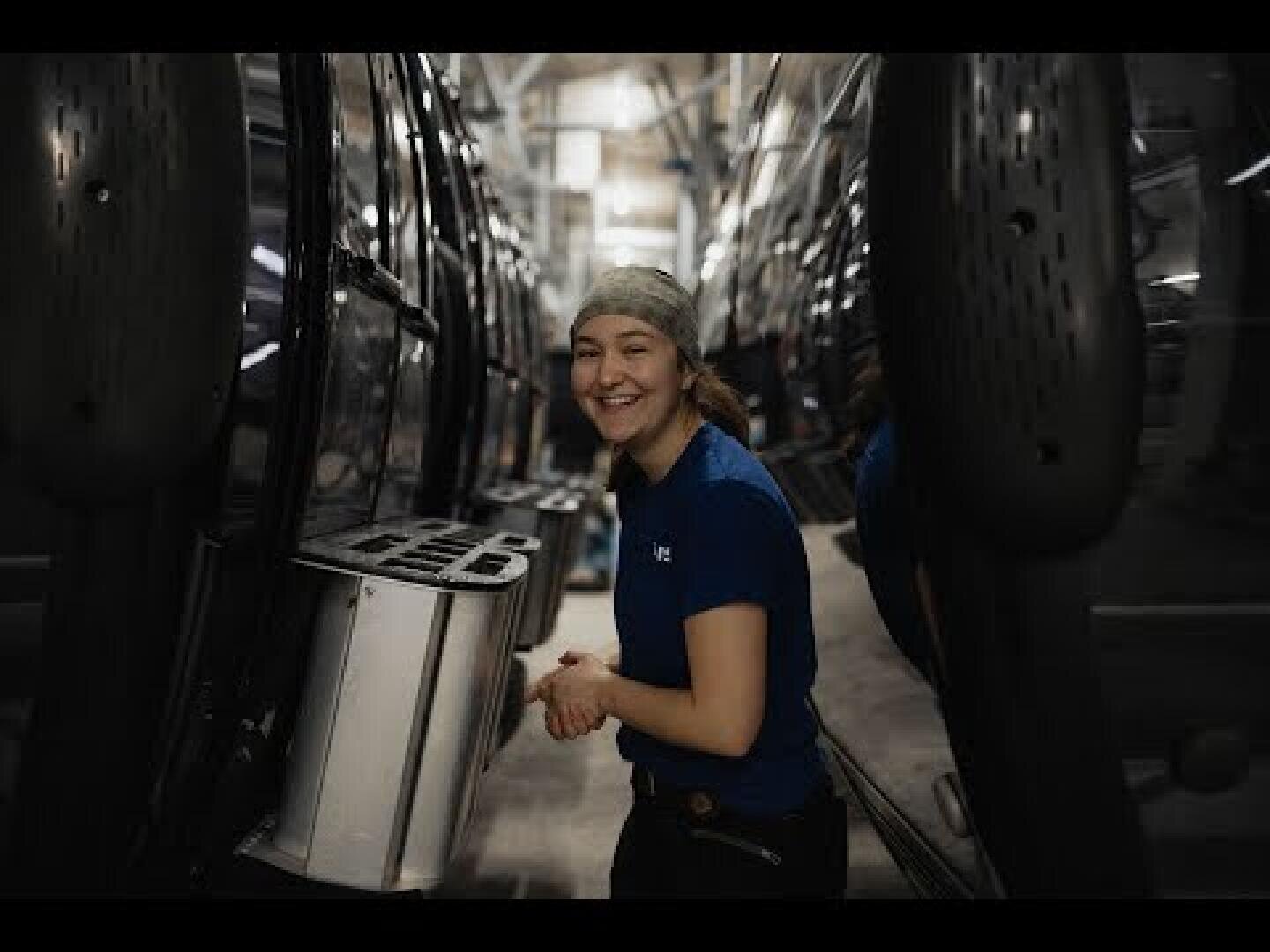 Eine Person, die ein blaues Hemd und ein Kopftuch trägt, lächelt, während sie zwischen zwei großen Fahrzeugen oder Maschinen in einer Fabrik oder einem Industriegebäude arbeitet.