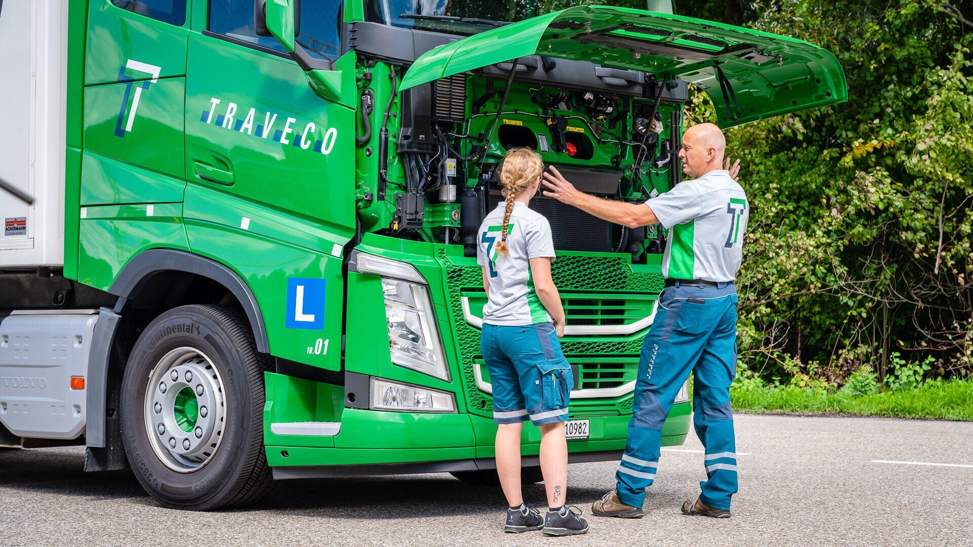 Zwei Personen in Uniform stehen neben einem grünen Lieferwagen mit geöffneter Motorhaube. Sie scheinen den Motor zu überprüfen oder darüber zu sprechen. Die Szene spielt im Freien in der Nähe einer Straße und inmitten von Grünflächen.