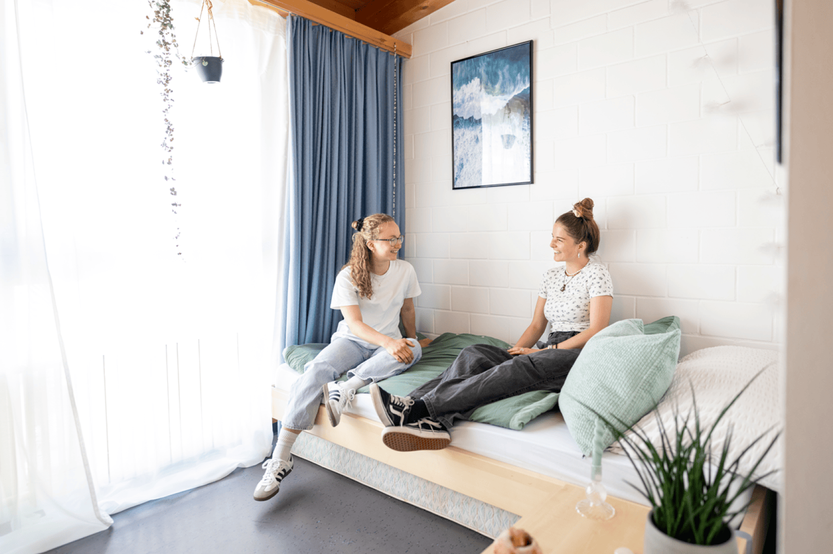 Zwei junge Frauen sitzen in einem hellen, modernen Zimmer mit großen Fenstern, blauen Vorhängen, Pflanzen und einem gerahmten Bild an einer weißen Backsteinwand auf einem sauber gemachten Bett und unterhalten sich.