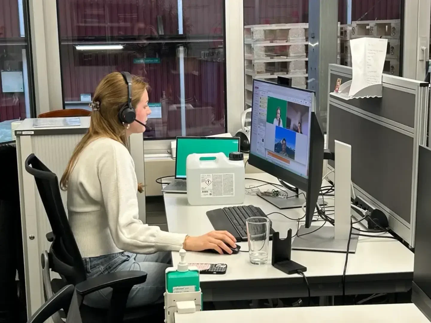 Eine Frau mit einem Headset sitzt an einem Schreibtisch und arbeitet an einem Computer, auf dessen Bildschirm ein Videoanruf zu sehen ist. Auf dem Schreibtisch stehen Büromaterial, ein Glas Wasser und eine grüne Flasche mit Desinfektionsmittel. Sie wirkt konzentriert.