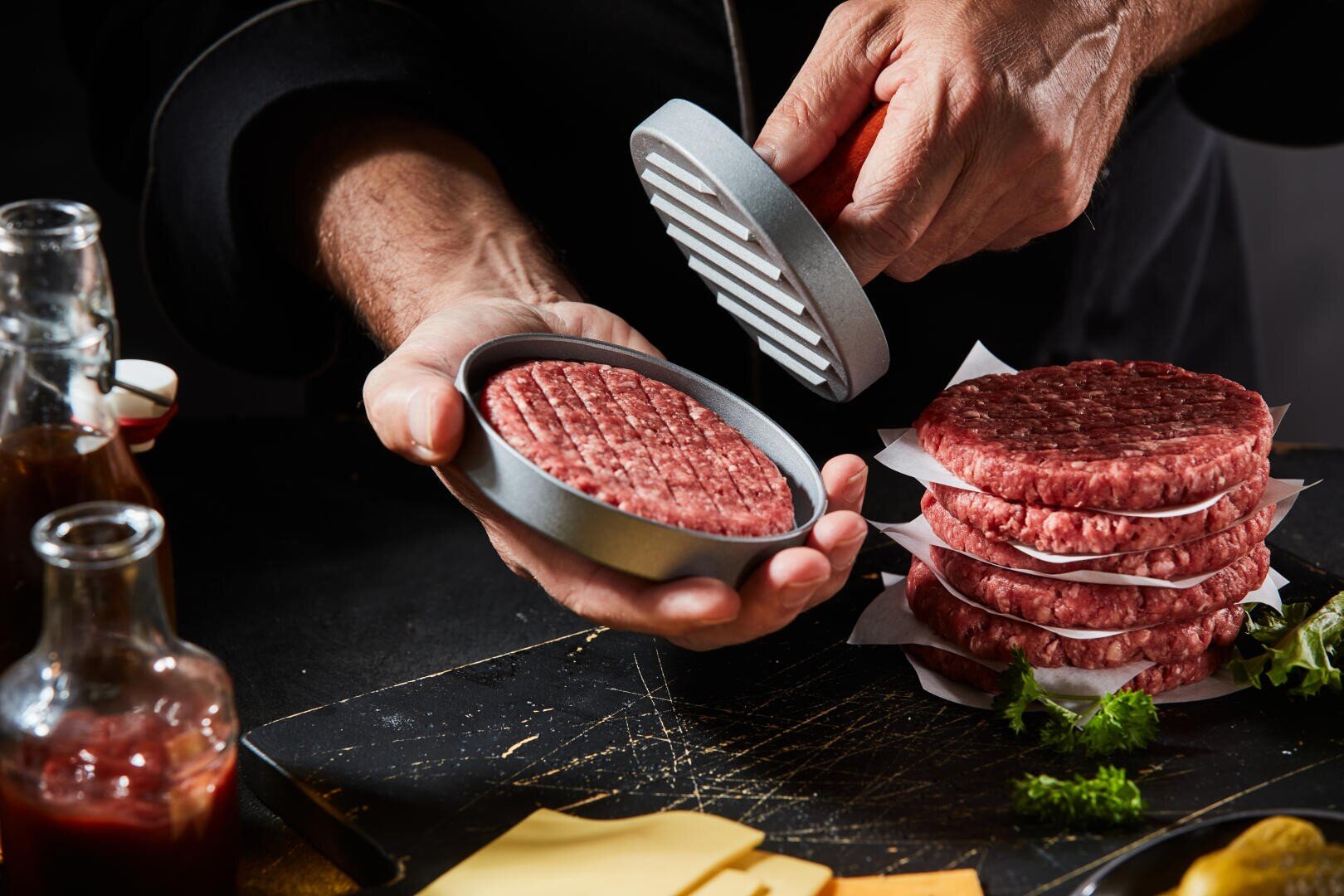Eine Person formt mit einer Metallpresse rohes Rinderhackfleisch zu Hamburger-Patties. Auf dem Tisch liegen mehrere gestapelte Patties, Käsescheiben und Soßenflaschen.