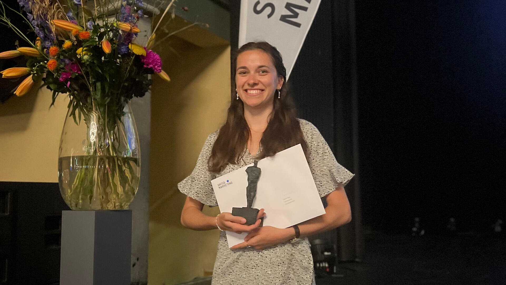 Eine Frau lächelt, während sie eine Auszeichnung und einen Umschlag in der Hand hält. Sie steht neben einer großen Vase mit bunten Blumen, mit einem Banner und einem dunklen Hintergrund hinter ihr.