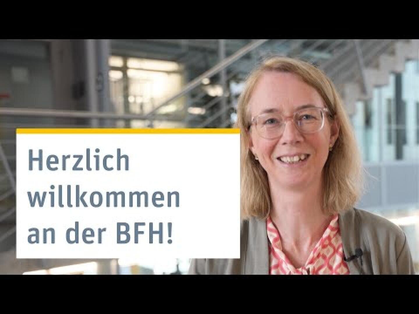 Eine lächelnde Frau mit Brille steht in einem Innenraum in der Nähe einer Treppe. Auf einem Schild neben ihr steht: "Herzlich willkommen an der BFH!".
