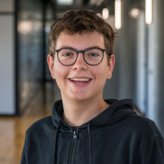 Ein junger Mann mit Brille, der in einem Büroflur lächelt.