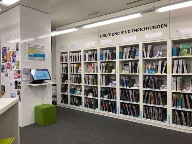 In der Bibliothek mit dem Titel Berufe und Studienrichtungen stehen Regale voller Bücher und Broschüren. Ein Computerarbeitsplatz mit Monitor und grünem Hocker befindet sich neben einer Wand, die mit Postern und Informationsblättern bedeckt ist.