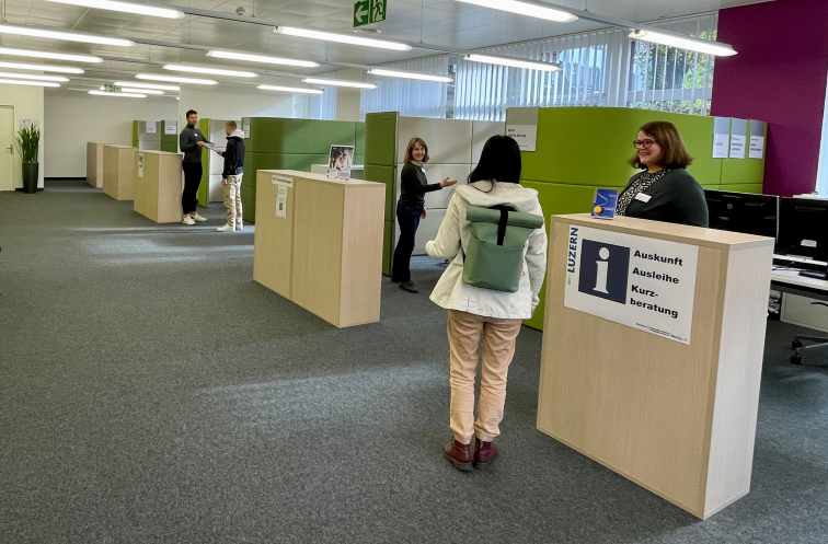 In einem modernen Büroraum mit grünen und lila Trennwänden stehen mehrere Personen an Serviceschaltern. Eine Frau mit grünem Rucksack unterhält sich mit einem lächelnden Mitarbeiter an einem Informationsschalter mit der Aufschrift Auskunft und Luzern.