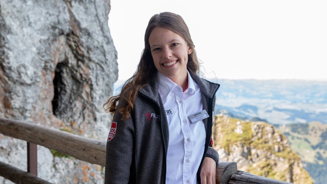 Eine junge Frau in grauer Jacke und wei&szlig;em Hemd l&auml;chelt, w&auml;hrend sie an einem Holzgel&auml;nder steht, mit felsigen Klippen und einer malerischen Landschaft im Hintergrund.
