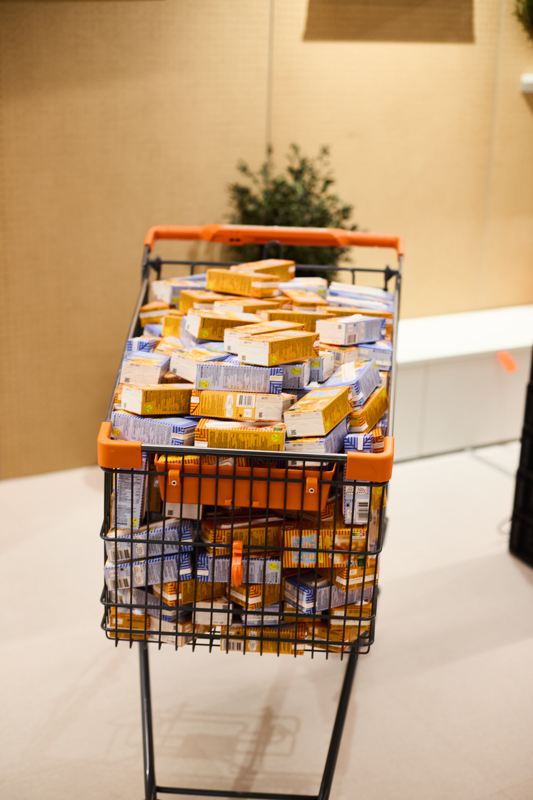 Ein Einkaufswagen, der bis oben hin mit zahlreichen kleinen, meist orangefarbenen und blauen Kartons gefüllt ist, in einem gut beleuchteten Innenraum.