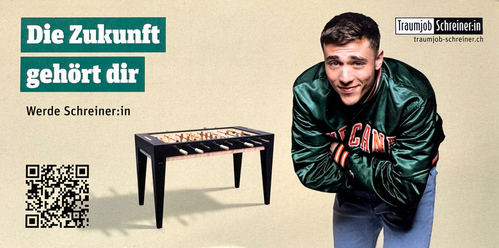 Ein junger Mann in einer grünen Jacke lehnt sich lächelnd neben einem Kickertisch nach vorne. Der Text lautet "Die Zukunft gehört dir. Werde Schreiner:in." mit einem QR-Code und dem Logo von traumjob-schreiner.ch.