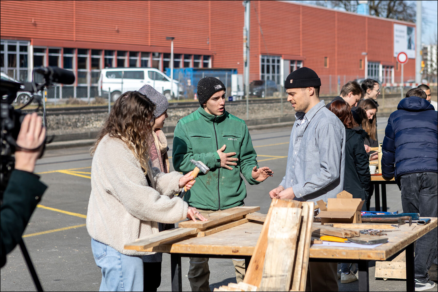 Mehrere junge Menschen stehen im Freien an Werkbänken und arbeiten mit Holz. Eine Person hält einen Hammer, während eine andere etwas erklärt. Im Hintergrund sind weitere Teilnehmende und ein rotes Industriegebäude zu sehen.