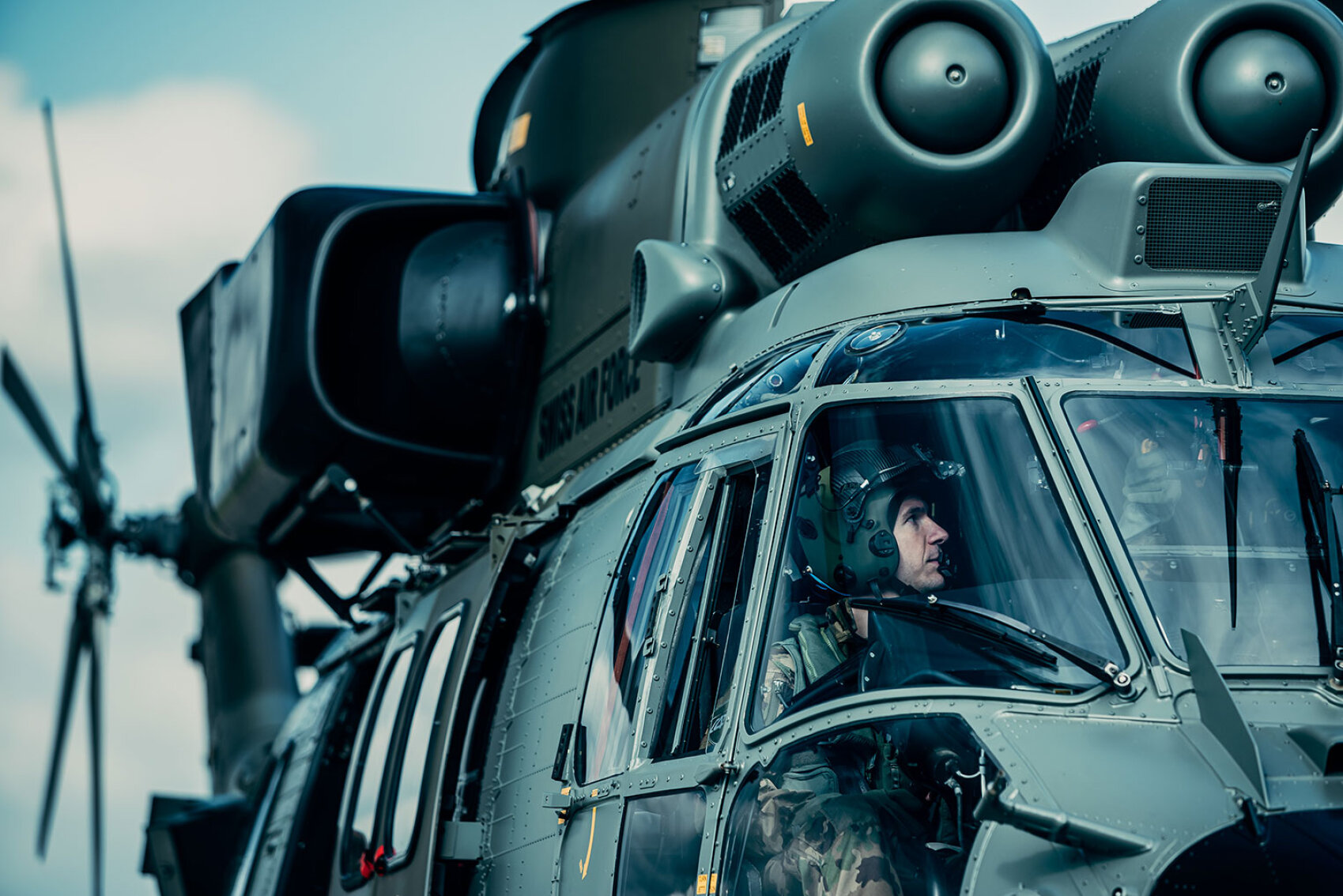 Ein Militärpilot mit Helm sitzt im Cockpit eines grünen Militärhubschraubers, wobei die Rotoren und äußeren Details des Flugzeugs deutlich sichtbar sind.