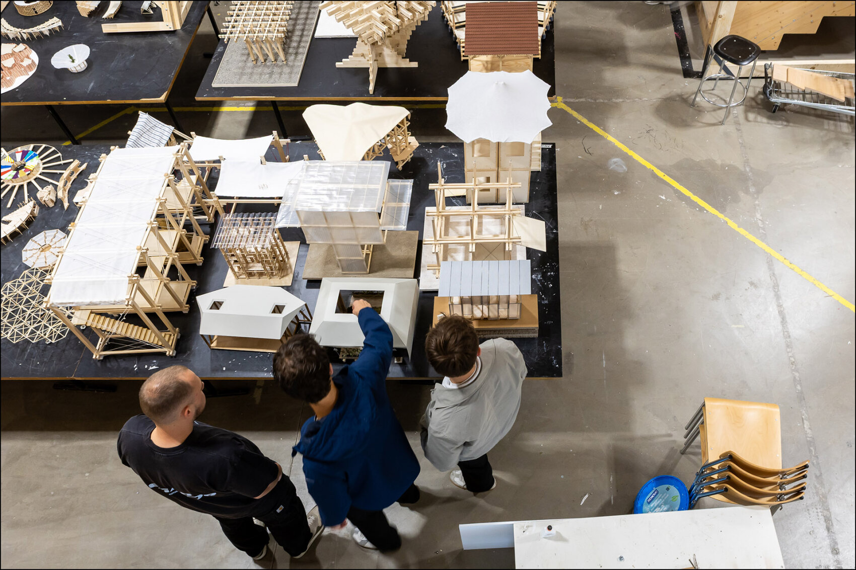 Drei Personen betrachten auf einem grossen Tisch mehrere detailreiche Architekturmodelle aus Holz und Karton. Die Modelle zeigen verschiedene Baukonstruktionen und stehen in einem Werkraum mit Betonboden.