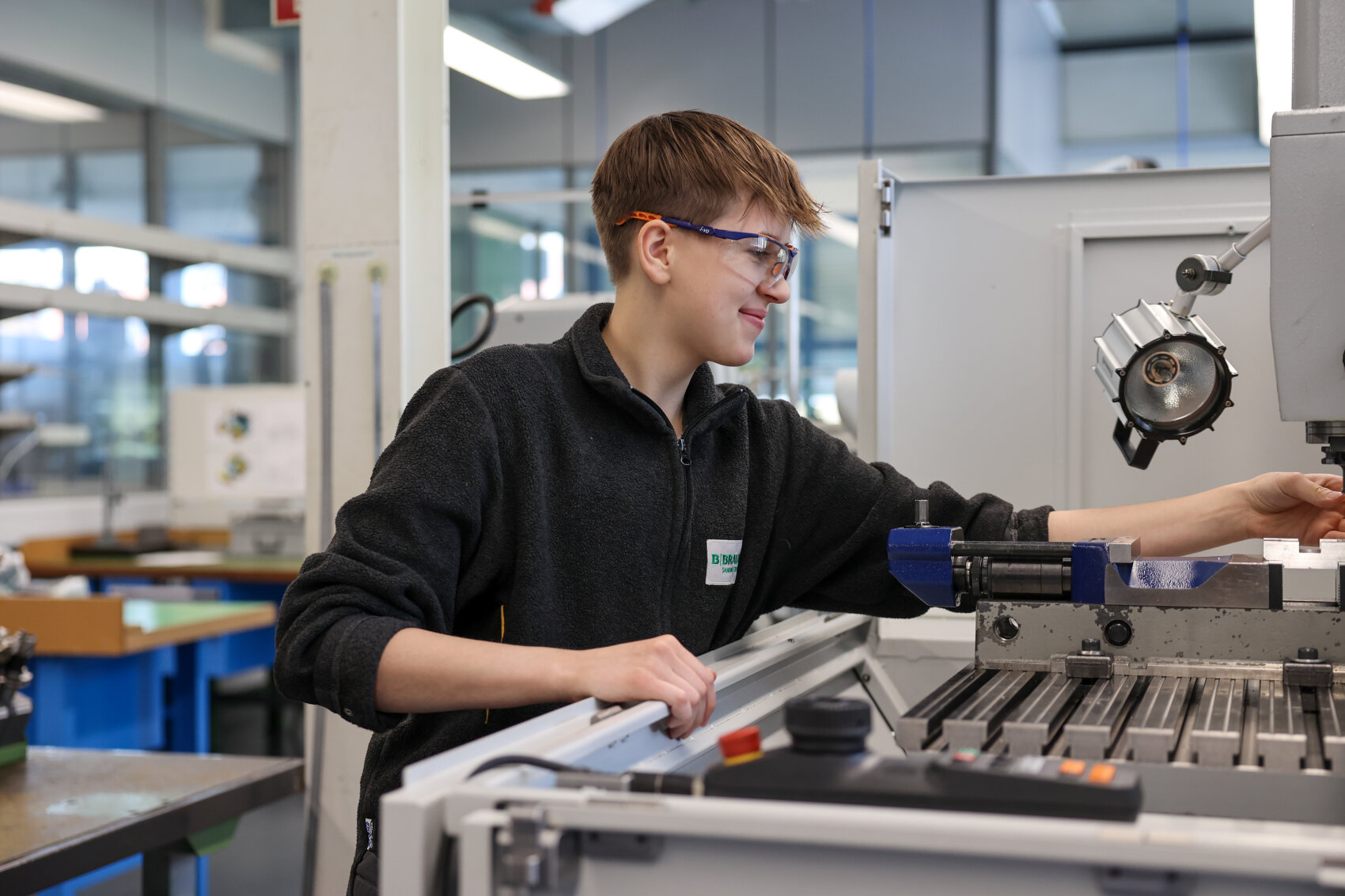 Ein junger Mann mit Schutzbrille bedient in einer hellen, modernen Werkstatt eine Maschine. Er konzentriert sich darauf, mit einer Hand ein Bauteil einzustellen, während er die Geräte betrachtet und lächelt.