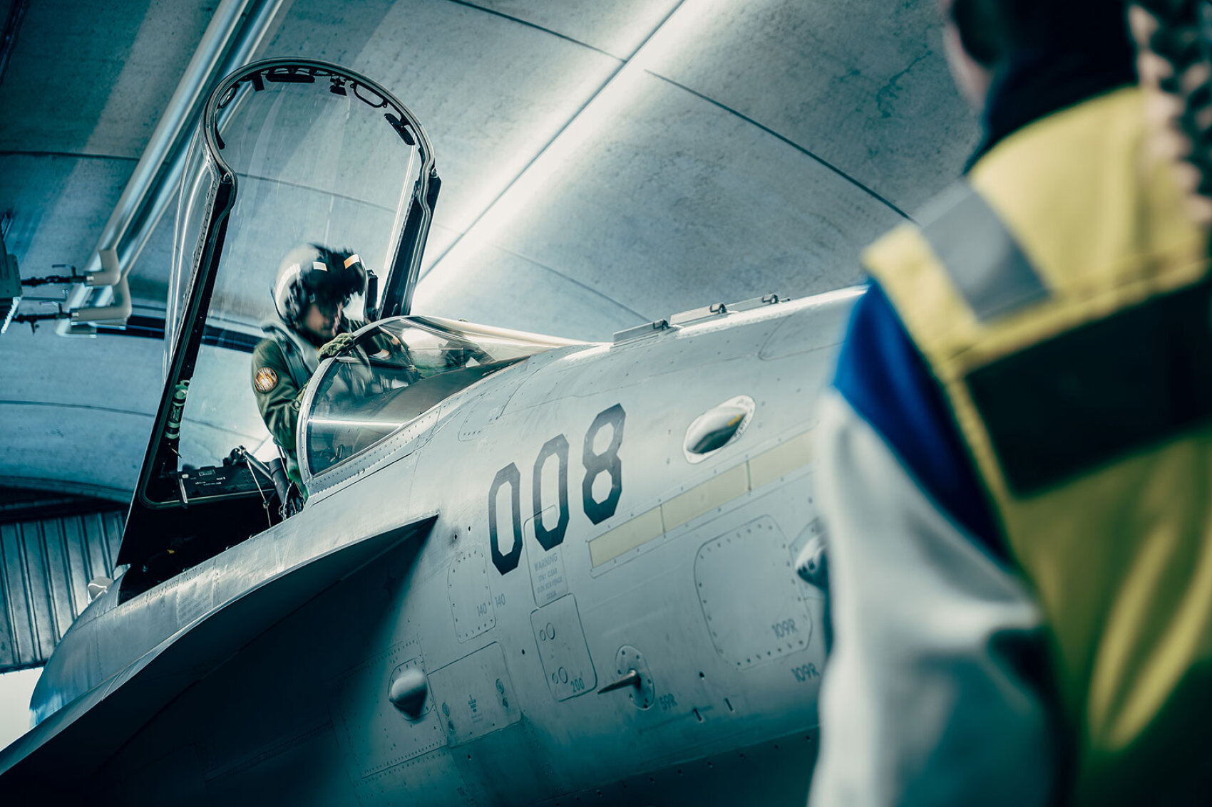 Ein Pilot sitzt im Cockpit eines Kampfflugzeugs mit der Nummer 008, dessen Kabinenhaube in einem Hangar geöffnet ist. Eine Person in einer reflektierenden Weste steht in der Nähe, teilweise sichtbar im Vordergrund.