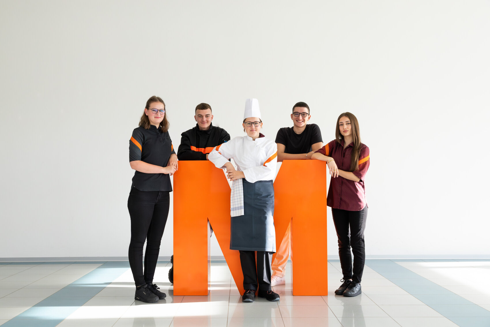Fünf junge Erwachsene stehen in einem hellen Raum um einen großen orangefarbenen Buchstaben M herum; einer trägt eine Kochuniform, die anderen sind in schwarzer oder kastanienbrauner Arbeitskleidung gekleidet und lächeln in die Kamera.