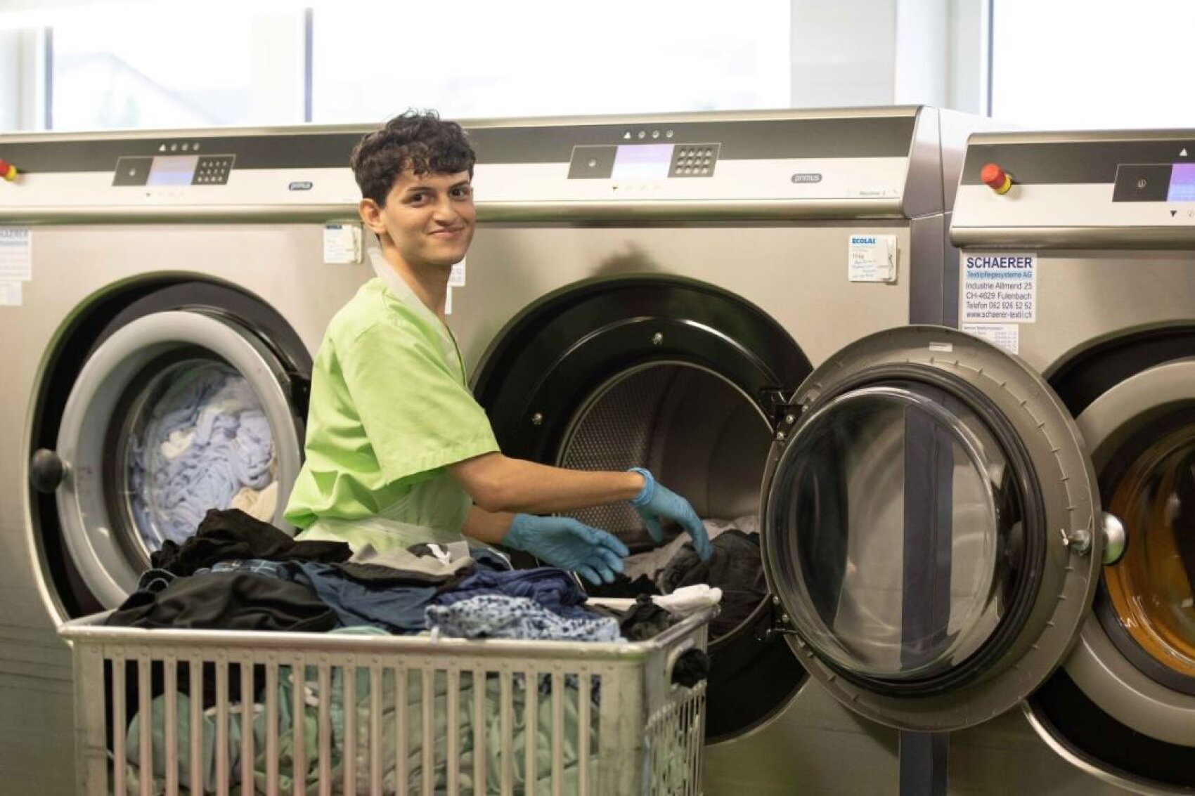 Eine Person in grüner Uniform und blauen Handschuhen lächelt, während sie in einem Waschsalon Wäsche in eine große Waschmaschine einlegt, vor der ein Wagen voller Wäsche steht.