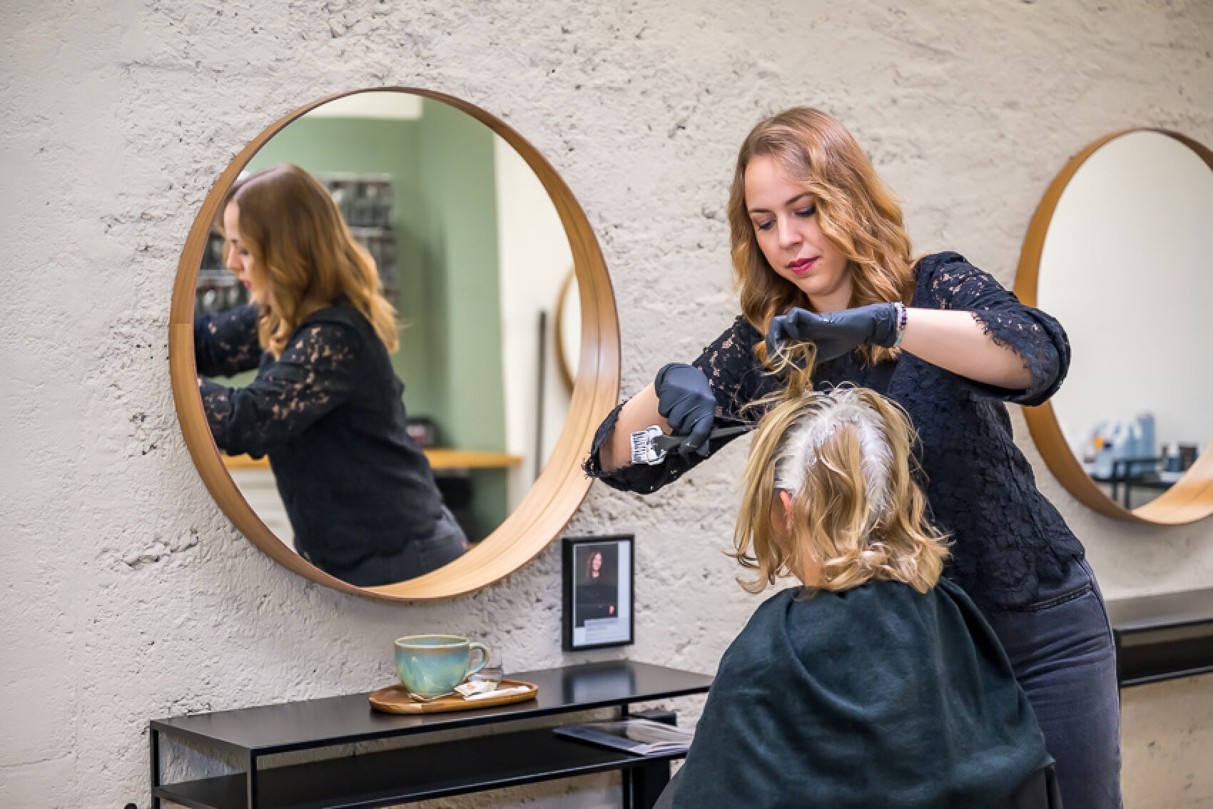 Eine Friseurin in schwarzer Kleidung und Handschuhen färbt einer sitzenden Kundin in einem modernen Salon mit runden Spiegeln und einer Tasse auf dem Tresen die Haare.