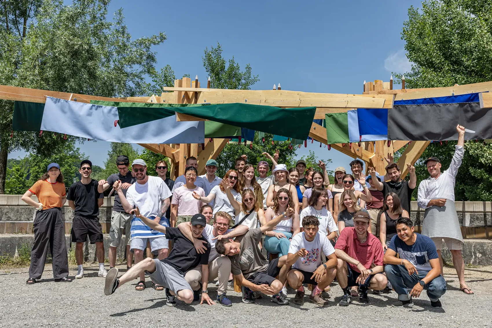 Eine grosse Gruppe junger Menschen posiert lachend vor einer Holzkonstruktion im Freien, über der bunte Stoffbahnen hängen. Die Sonne scheint, und die Stimmung ist fröhlich und ausgelassen.
