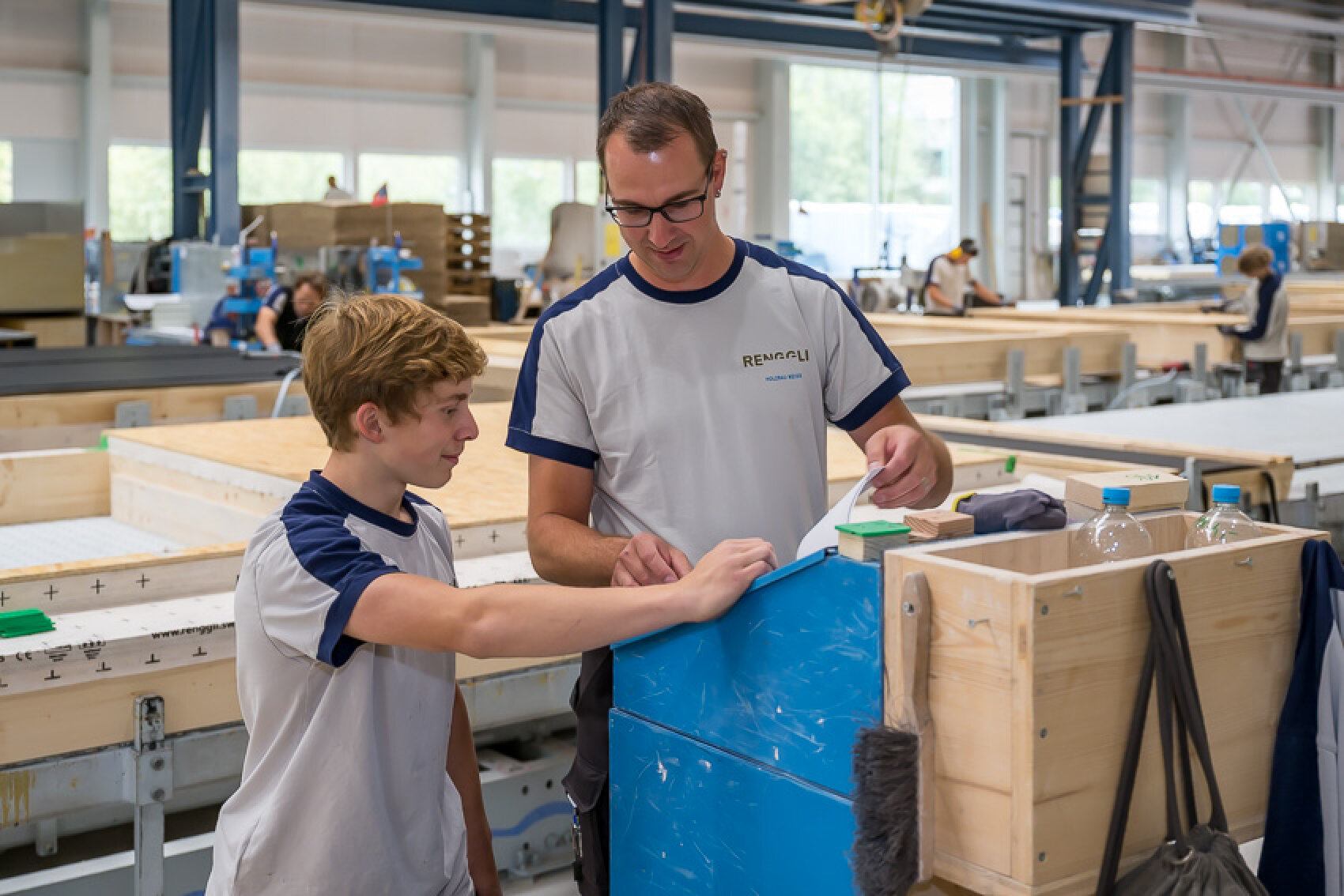 Ein Mann und ein Junge, beide in weißen und blauen Hemden, stehen zusammen in einer Fabrik, lächeln und diskutieren über etwas auf einer blauen Arbeitsfläche. Im Hintergrund sind andere Arbeiter und Geräte zu sehen.