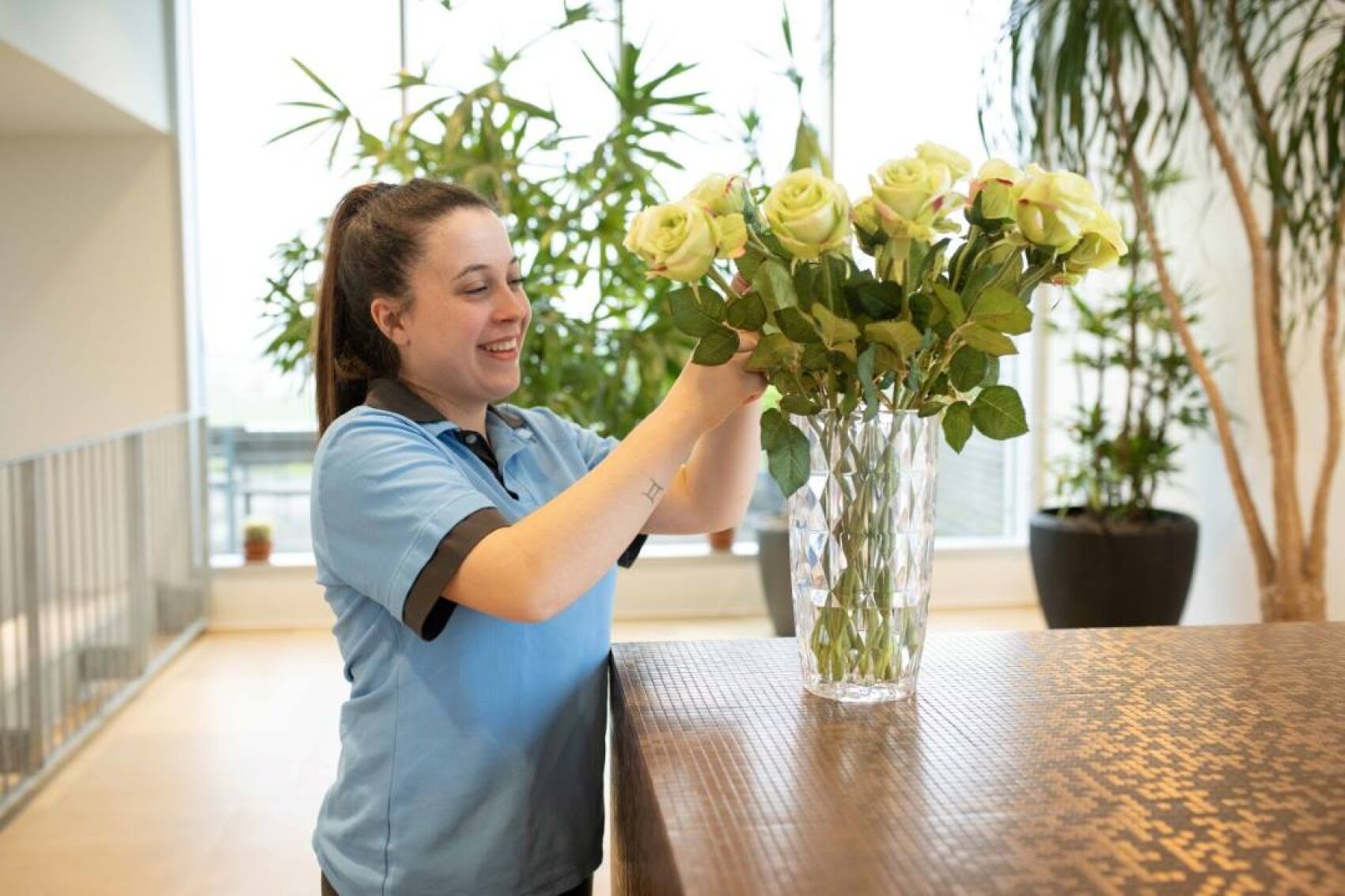 Eine Frau in blauer Uniform arrangiert lächelnd gelbe Rosen in einer Glasvase auf einer Arbeitsplatte, mit großen Grünpflanzen und Fenstern im hellen Hintergrund.