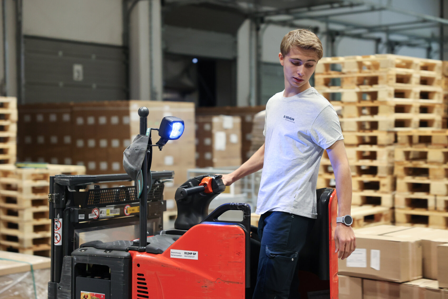 Eine junge Person bedient einen roten Palettenheber in einem Lagerhaus, umgeben von Stapeln von Holzpaletten und Kartons. Die Person trägt ein hellgraues Hemd und scheint sich auf das Manövrieren des Geräts zu konzentrieren.