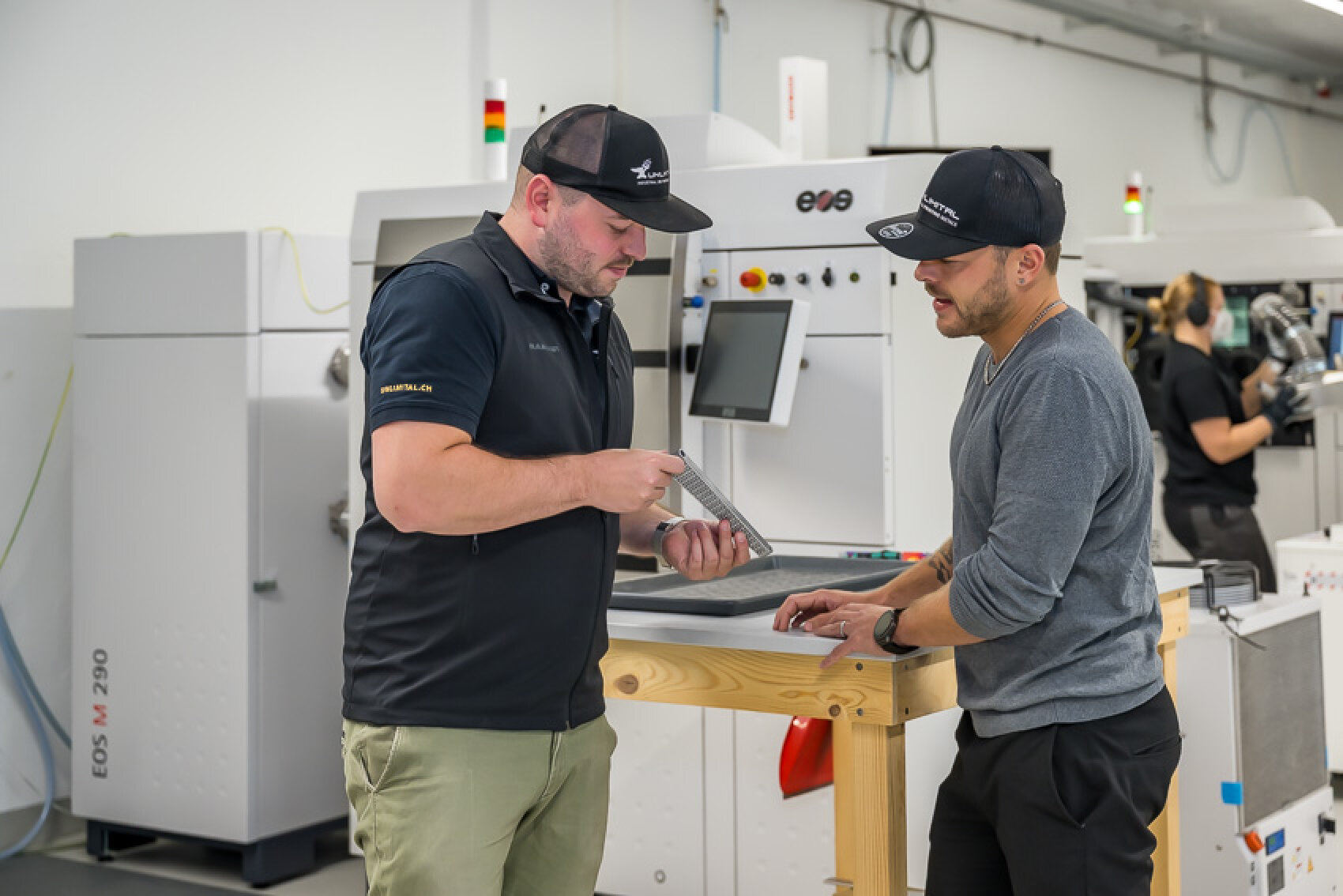 Zwei Männer in Arbeitsuniformen diskutieren in einem modernen Industrielabor über ein Metallteil. Sie stehen an einem Holztisch mit Maschinen und Computern im Hintergrund. Eine weitere Person bedient Geräte im Hintergrund.