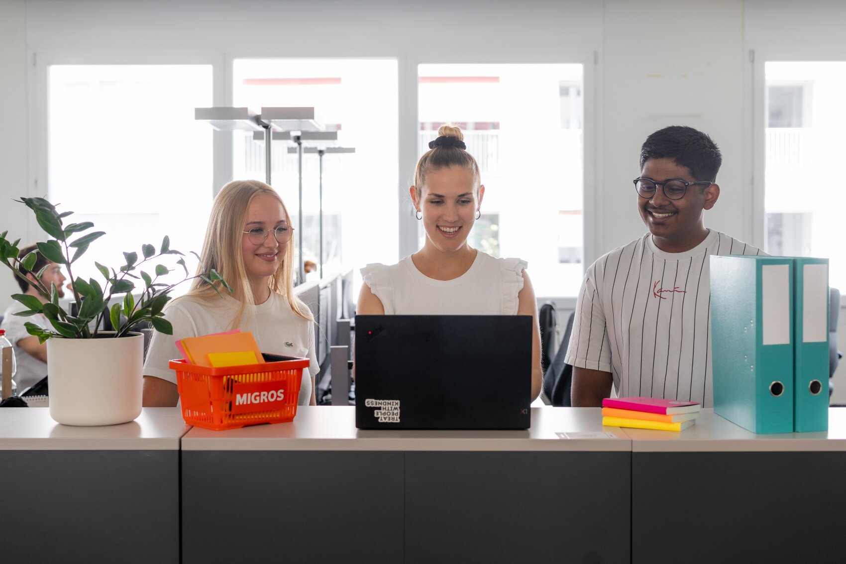 Drei junge Erwachsene, zwei Frauen und ein Mann, lächeln, während sie auf einen Laptop auf einem Bürotisch schauen. In der Nähe liegen bunte Mappen, Haftnotizen, eine Topfpflanze und ein orangefarbener Korb mit der Aufschrift "Migros". Im Hintergrund sind helle Fenster zu sehen.