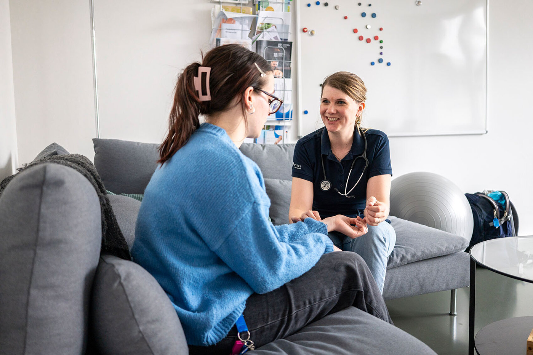 Eine medizinische Fachkraft mit Stethoskop unterhält sich lächelnd mit einem Patienten im blauen Pullover, der in einem hellen, gemütlichen Raum auf einer grauen Couch sitzt.