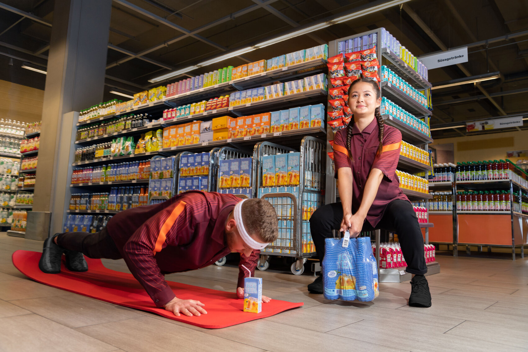 Zwei Supermarktangestellte in Uniform trainieren in einem Gang; einer macht Liegestütze über einer Saftpackung auf einer Matte, während der andere eine Packung Getränkeflaschen wie Gewichte hebt. Im Hintergrund sind Regale mit Getränken und Lebensmitteln zu sehen.