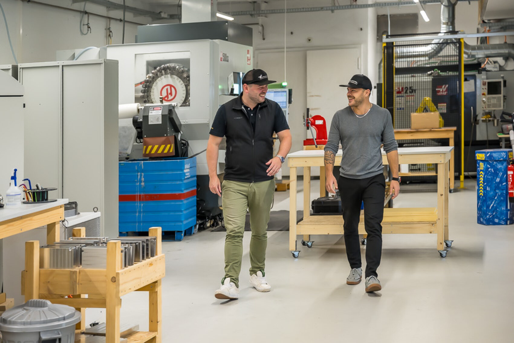 Zwei Männer in Freizeitkleidung und mit Mützen gehen und unterhalten sich in einer sauberen, modernen Industriehalle mit verschiedenen Maschinen, Werkzeugen und Werkbänken um sie herum.