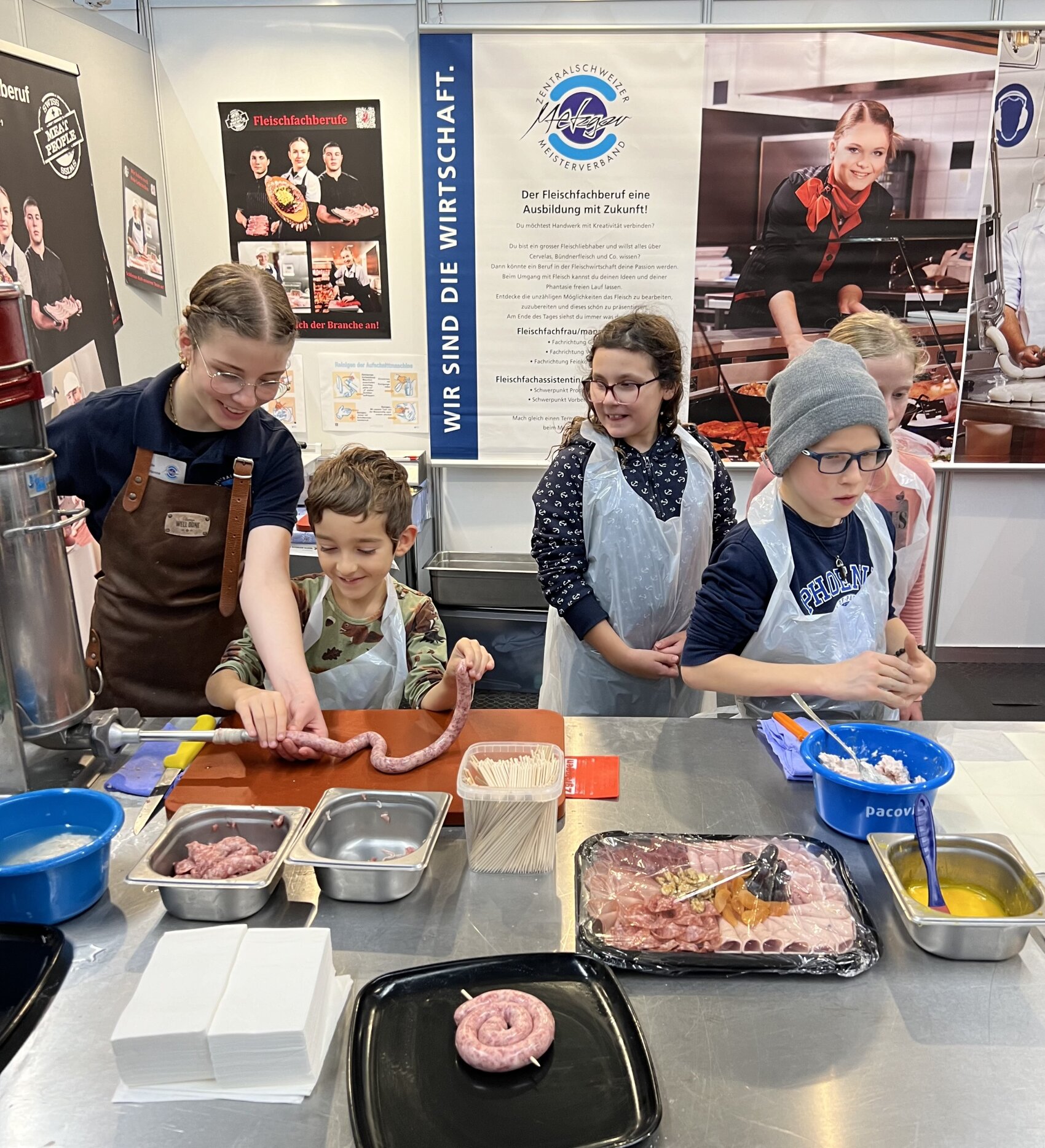Vier Kinder und ein Erwachsener stehen an einem Tisch und stellen Würste mit rohem Fleisch und Wursthüllen her. Sie tragen Schürzen in einer Küche oder einem Lebensmittelvorführbereich mit Postern und Kochgeräten im Hintergrund.